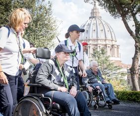 Im Hintergrund sind die vatikanischen Gärten zu sehen und im Vordergrund eine Pilgerin im Rollstuhl mit Malteser Helferinnen die gemeinsam einem Audioguide lauschen.
