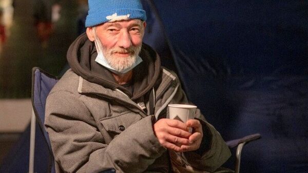 Ein älterer Mann mit hellblauer Mütze sitzt mit einem Becher auf einem Campingstuhl.