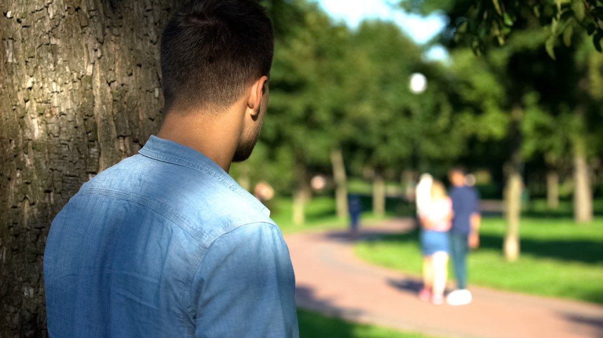Ein Mann hinter einem Baum beobachtet ein Pärchen im Hintergrund.