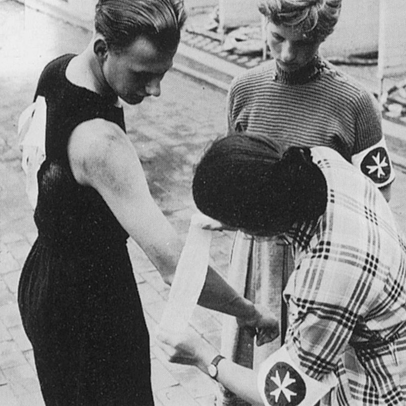 Ein Schwarzweißbild, auf dem eine Frau einem Sportler einen Verband anlegt, während eine andere Frau mit Armbinde des Malteser Hilfsdienstes dabei zusieht.