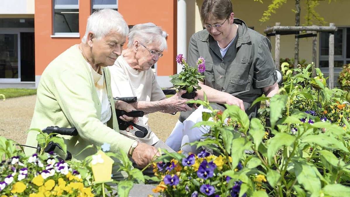 Zwei ältere Frauen und eine jüngere Frau bepflanzen ein Blumenbeet vor einem Gebäude.