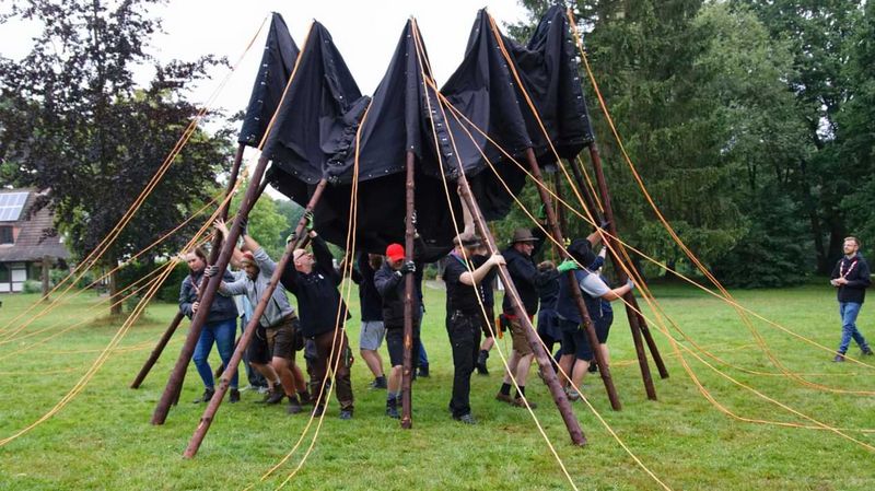 Eine Gruppe Menschen baut gemeinsam ein großes Zelt mit schwarzer Plane auf