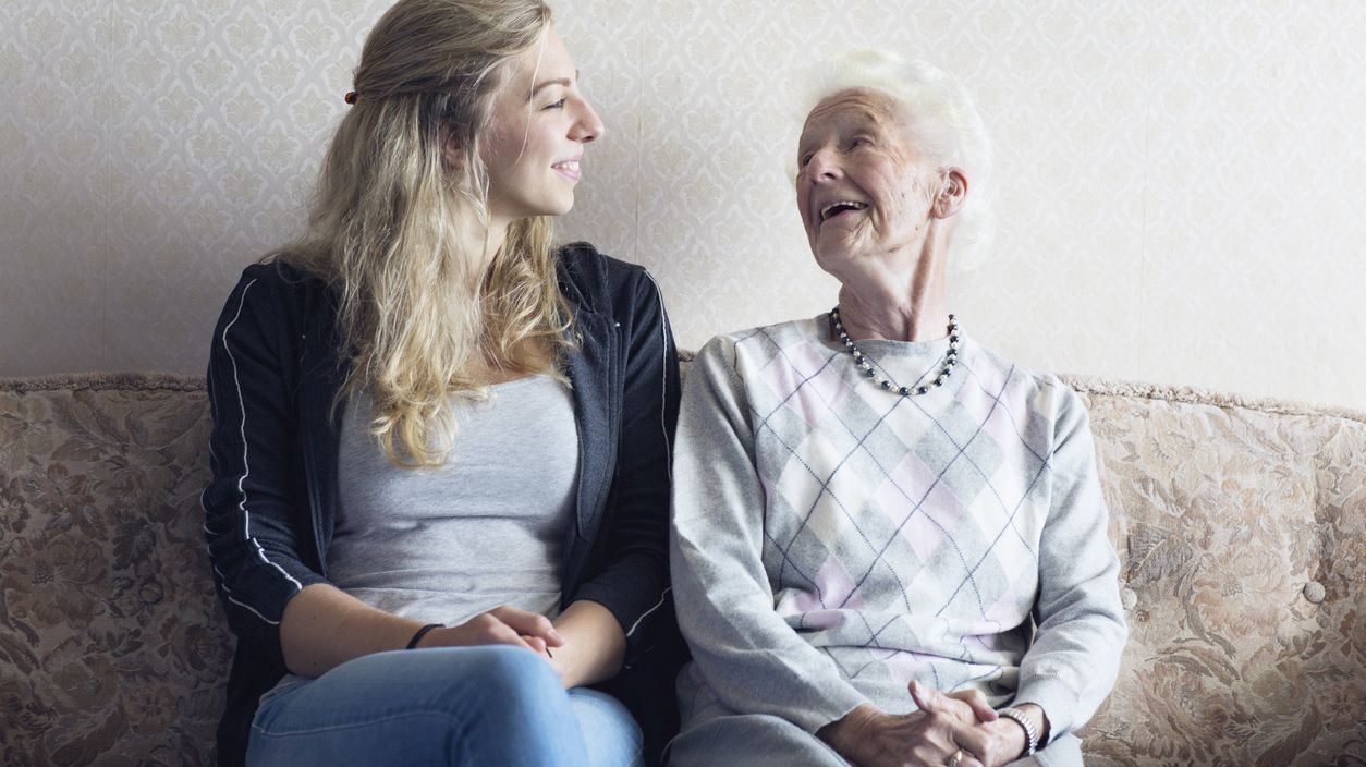 Junge Frau und Seniorin lächeln sich sitzend auf einem Sofa an.