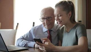 Junge Frau erklärt älterem Mann Smartphone und Laptop.
