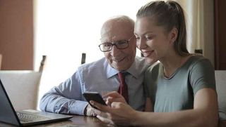 Junge Frau erklärt älterem Mann Smartphone und Laptop.