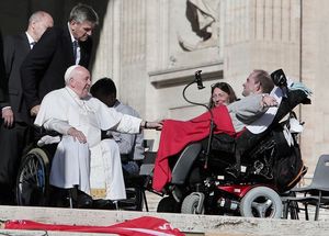 Papst Franziskus reicht einem Menschen im Rollstuhl die Hand. 