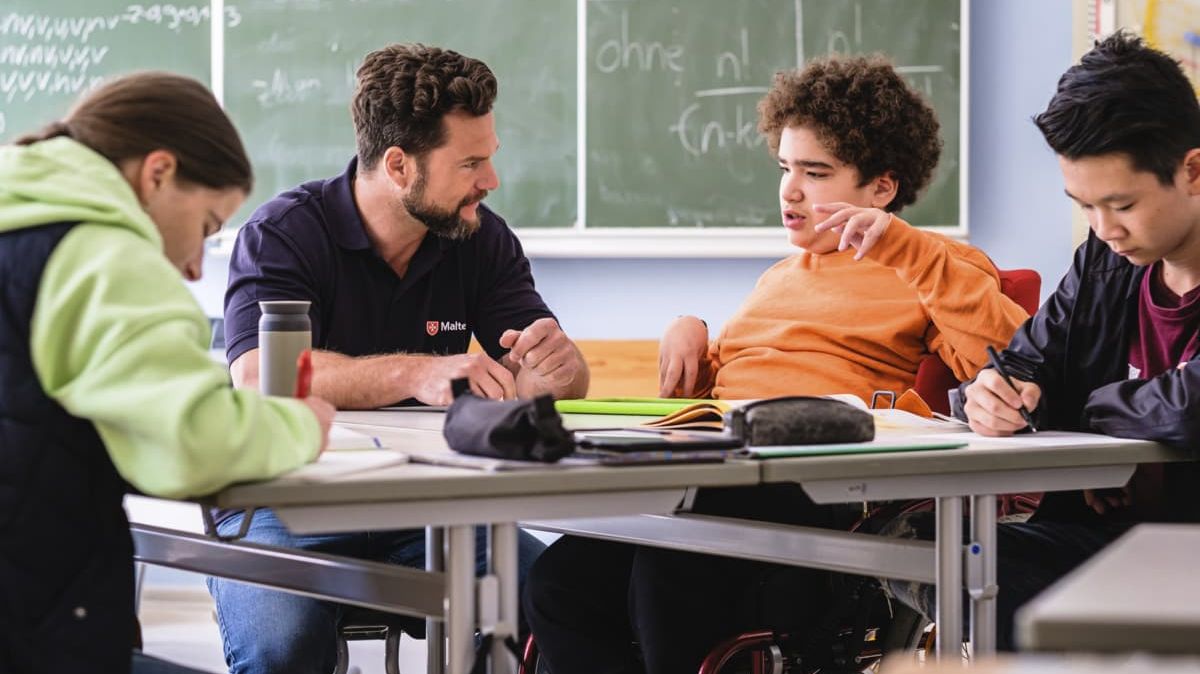 Malteser Schulbegleiter sitzt mit Schülern im Klassenzimmer.