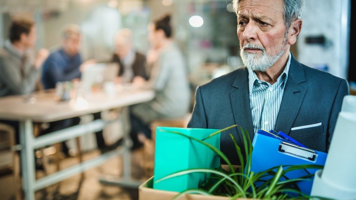 Ein älterer Mann mit ernstem Gesicht mit einem Karton voller Büroutensilien.