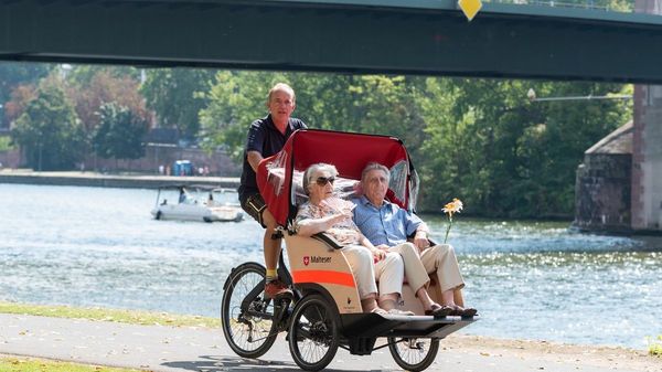 Älteres Paar sitzt in einer Rikscha, die von einem Mann gefahren wird, im Hintergrund eine Brücke und ein Boot.
