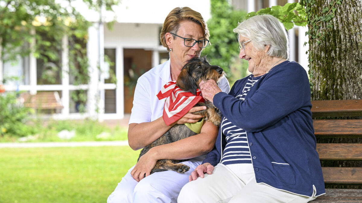 Eine lächelnde Seniorin sitzt zusammen mit einer Angehörigen, die einen Dackel auf dem Arm hält, auf einer Parkbank und streichelt den Hund.
