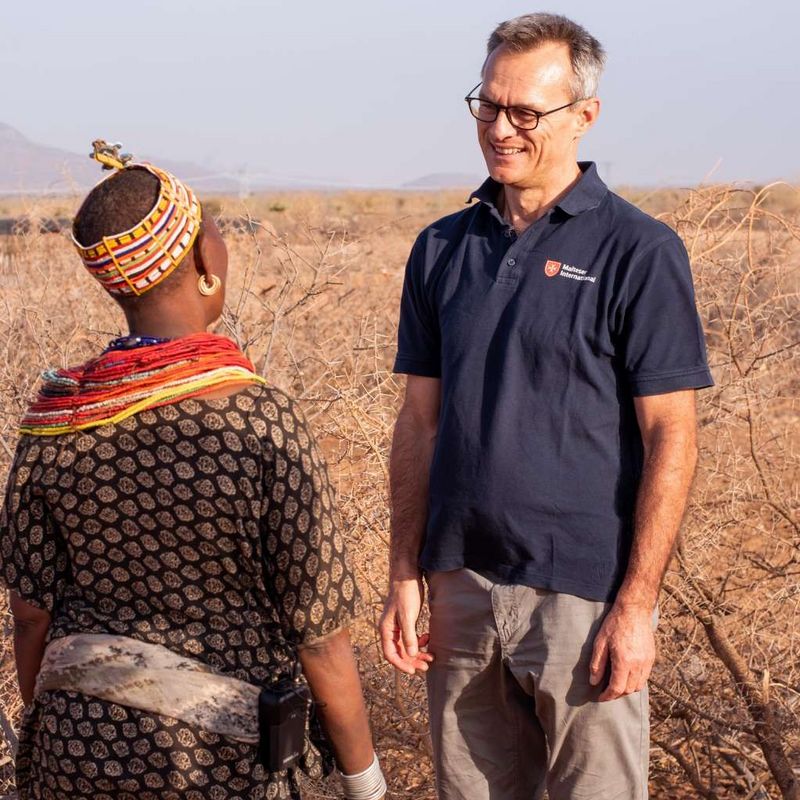 Ein lächelnder Mann mit Brille steht einer Person mit afrikanischem Kopfschmuck in einer Steppenlandschaft gegenüber