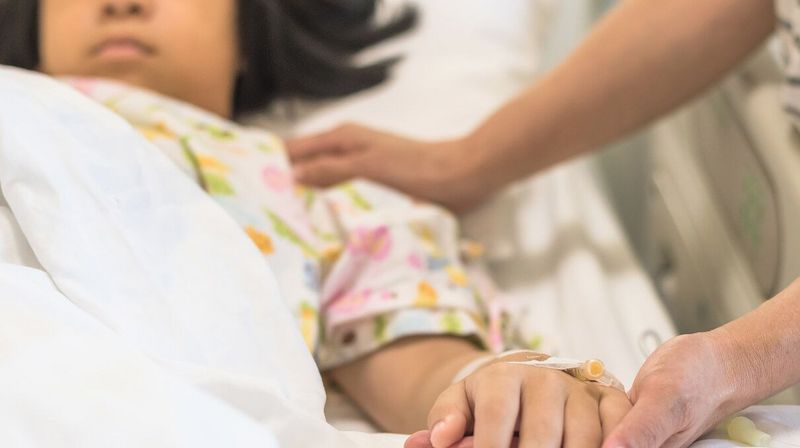 Eine Person hält die Hand eines Mädchens, das in einem Krankenhausbett liegt.