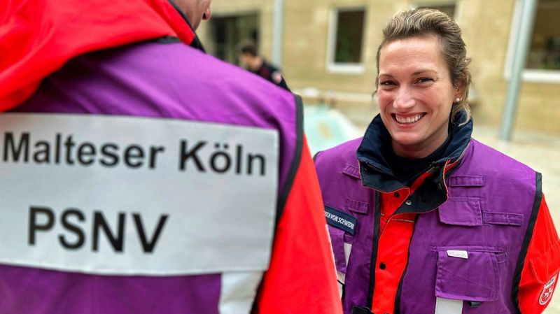 Eine lachende Frau in Einsatzkleidung und lila Weste. Im Vordergrund ist eine lila Weste mit der Aufschrift: „Malteser Köln PSNV“ zu sehen.