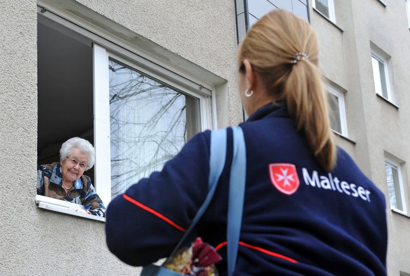 Eine Malteser Mitarbeiterin steht mit Einkäufen unter einem Fenster und unterhält sich mit einer älteren Dame.
