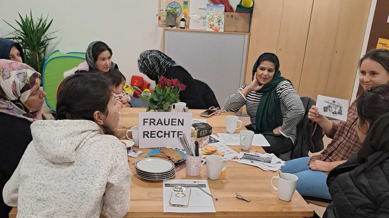 Frauen mit und ohne Kopftuch sitzen an einem Tisch, auf dem ein Schild mit der Aufschrift „Frauenrechte“ steht. 