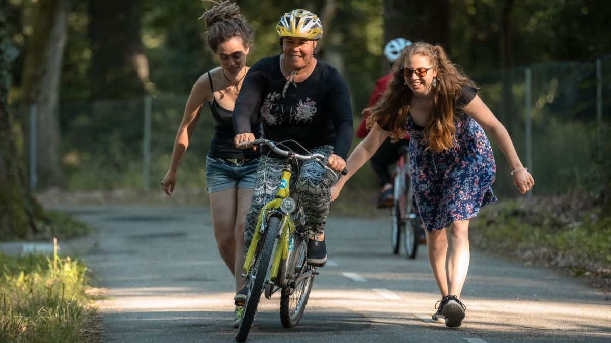 Zwei junge Frauen schieben eine Frau auf einem Fahrrad an.