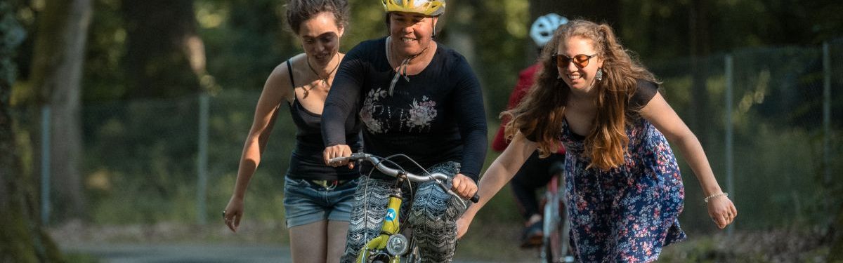 Zwei junge Frauen schieben eine Frau auf einem Fahrrad an.