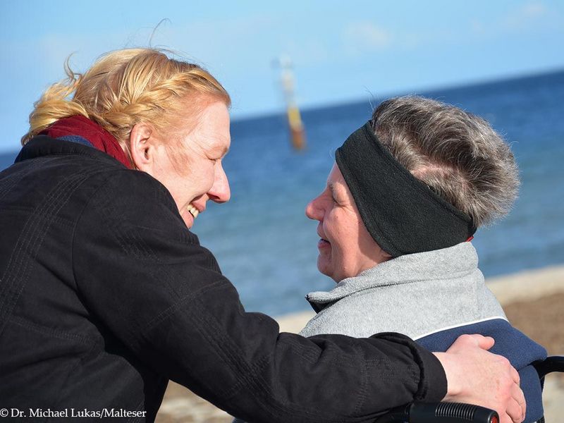 Eine Mitarbeiterin der Malteser und eine Frau im Rollstuhl lächeln sich an. Sie sind am Meer.
