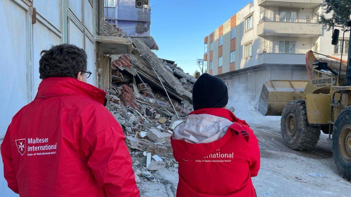 Zwei Menschen mit roten Jacken vor einem zerstörten Haus.