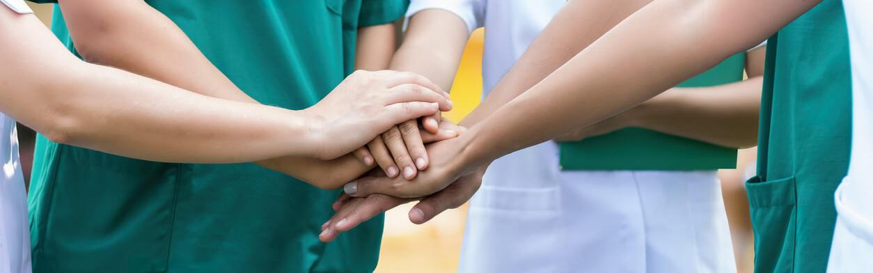 Pflegekräfte stehen im Kreis zusammen und symbolisieren Gemeinschaft, in dem sie ihre Hände zusammen aufeinanderlegen.