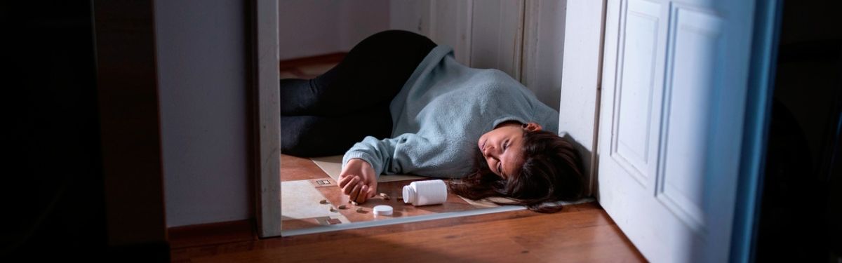 Eine bewusstlose Frau liegt auf dem Boden neben einer geöffneten Pillendose.