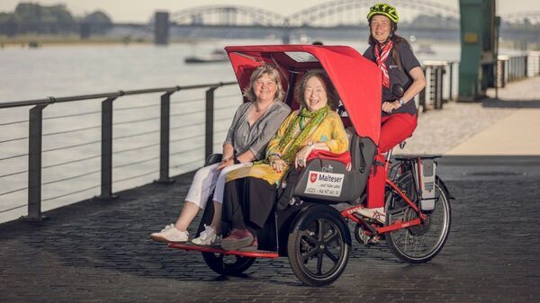 Eine lächelnde Frau mit Fahrradhelm befördert zwei ältere Frauen mit einer E-Rikscha.