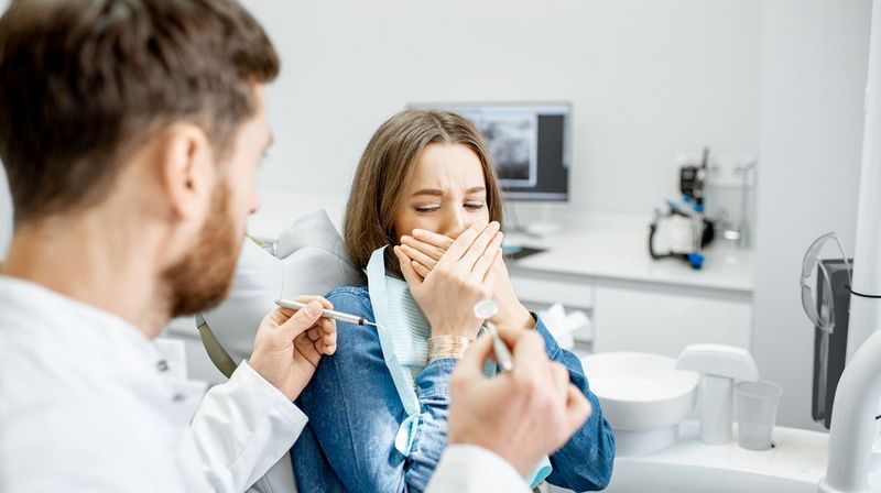 Frau auf dem Behandlungsstuhl beim Zahnarzt hält sich den Mund zu und hat Angst