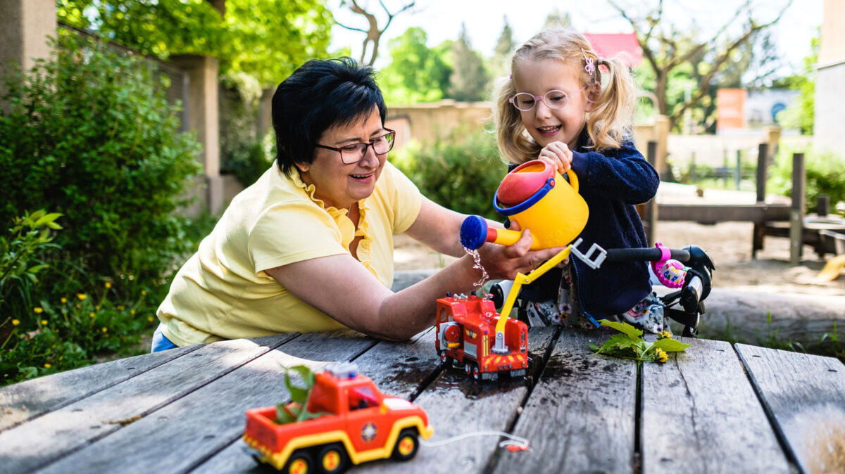 Eine Mutter spielt mit ihrer kleinen Tochter mit Plastikspielzeug auf einem Spielplatz-Outdoor-Holztisch.