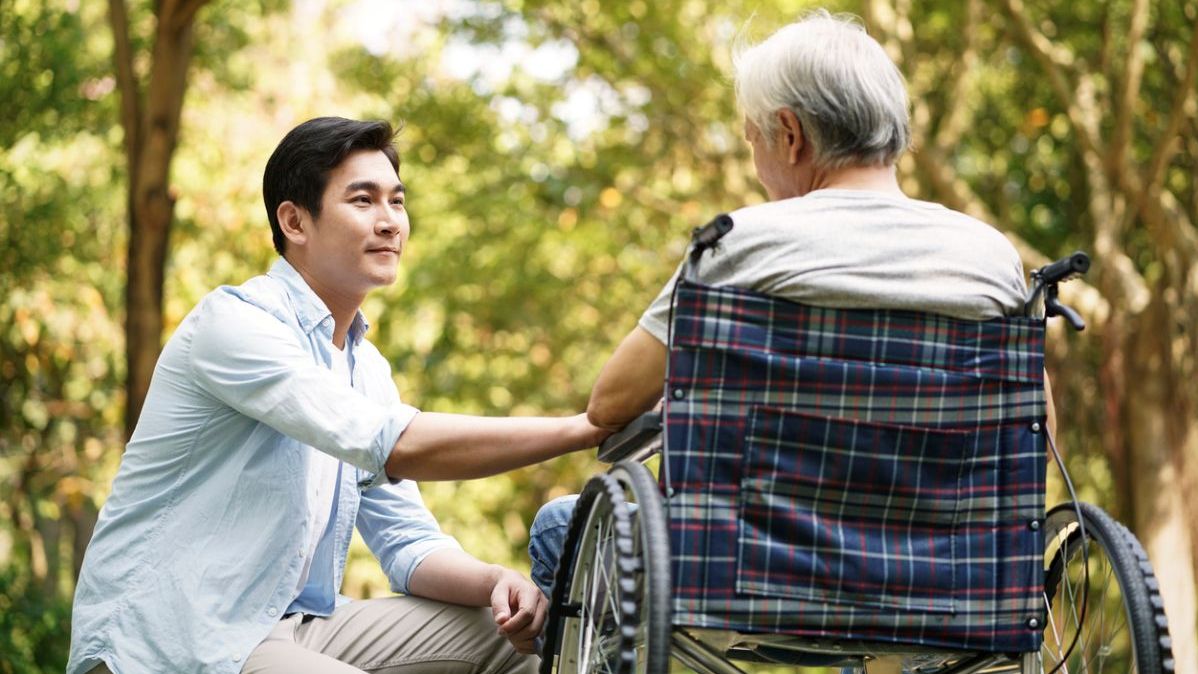 Junger Mann kniet vor einer älteren Person im Rollstuhl