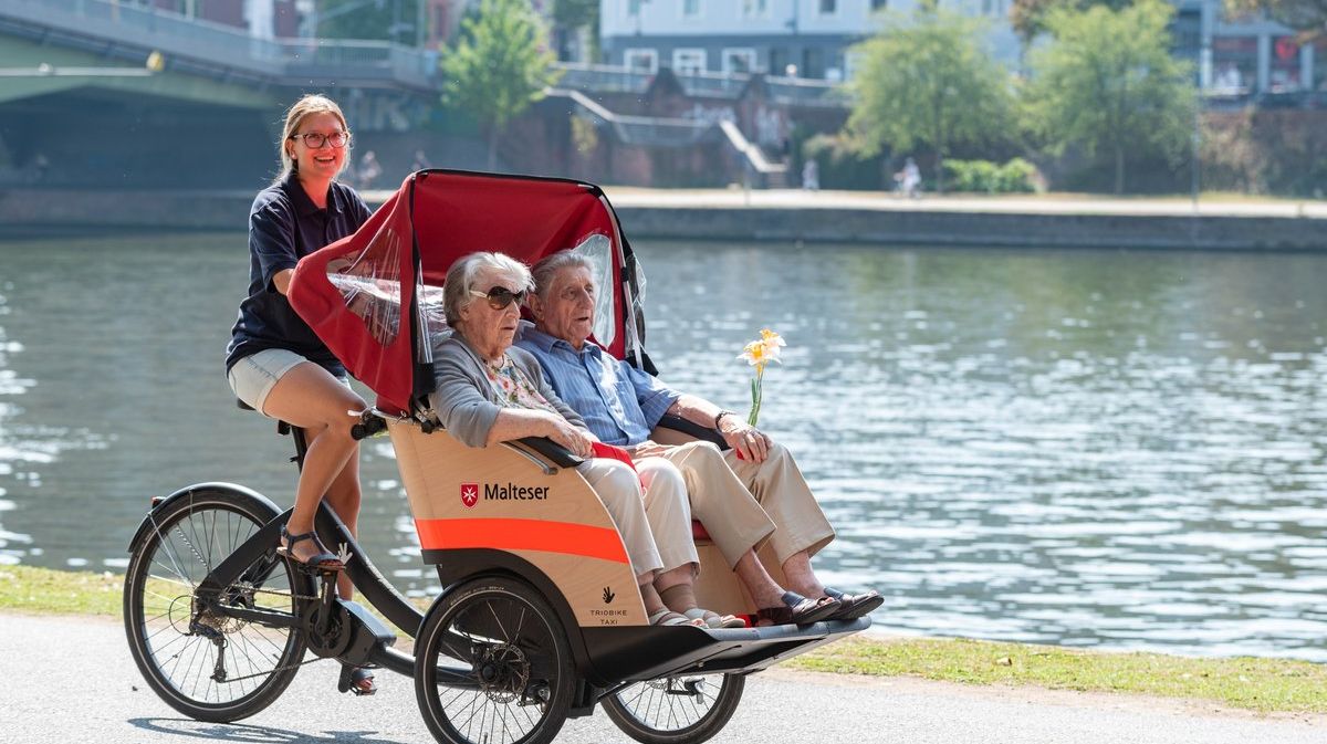 Älteres Paar sitzt in einer Rikscha, die von einer Frau gefahren wird, im Hintergrund ein Fluss.