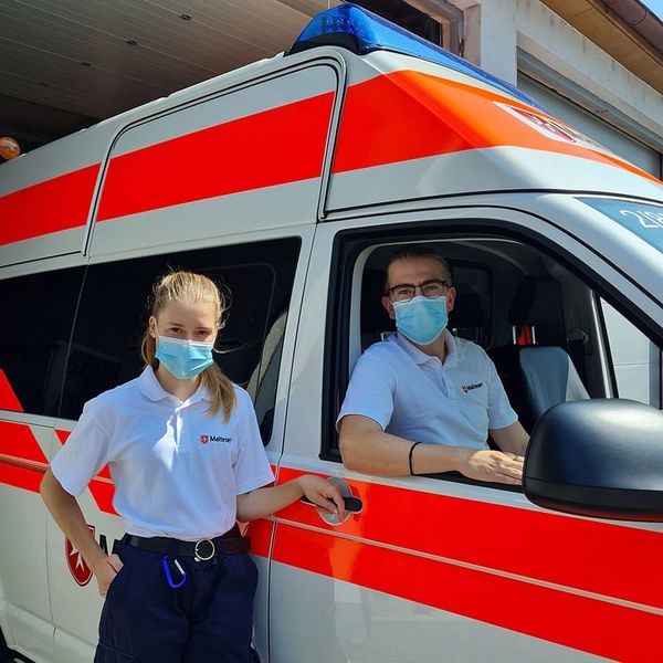 Ein Mann mit Mund-Nasen-Schutz in einem Krankenwagen und eine Frau mit Mund-Nasen-Schutz vor dem Wagen.