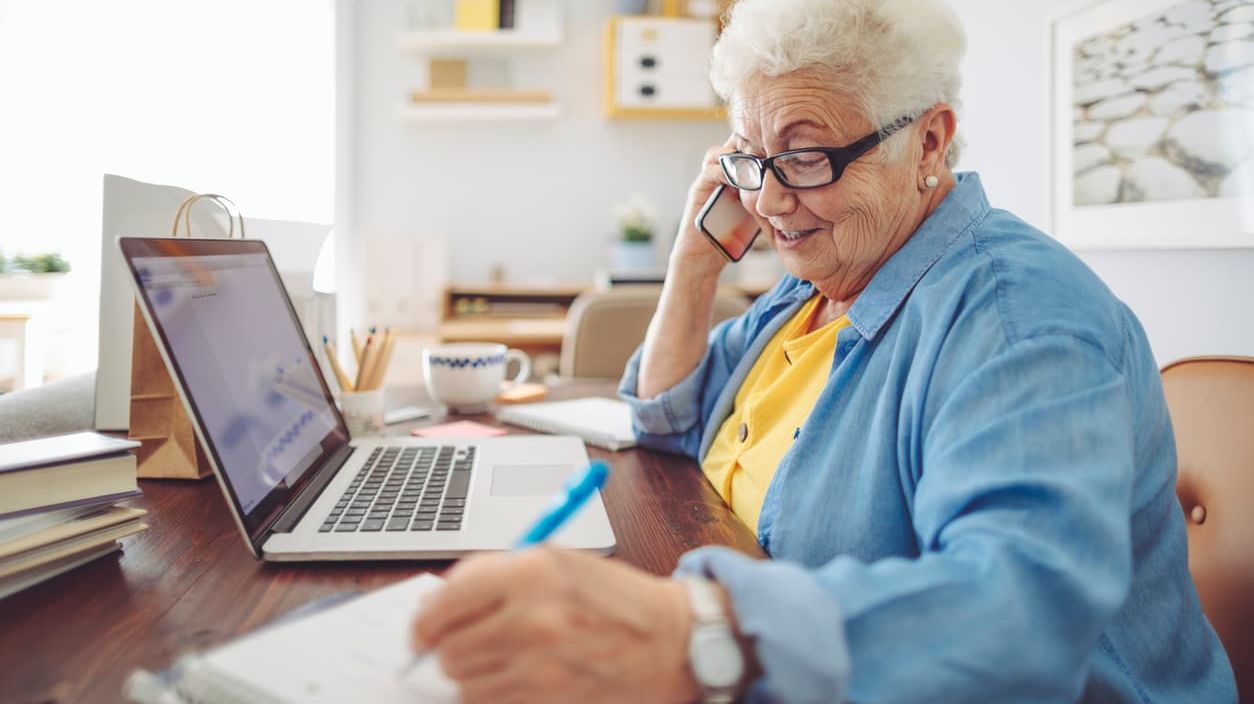 Ältere Frau sitzt telefonierend vor einem Laptop und notiert etwas auf einem Notizblock.