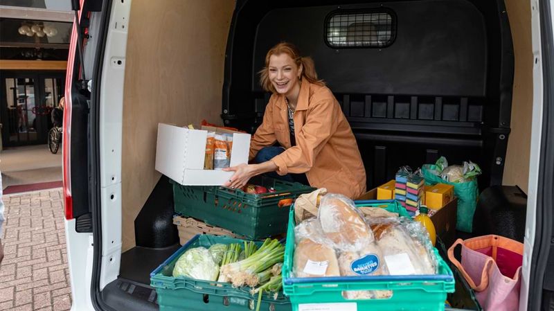 Eine lächelnde Frau kniet in einem Kleintransporter, in dem Lebensmittelkisten stehen.