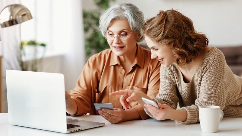 Jüngere Frau hilft ältere Frau am Rechner beim Online-Banking.