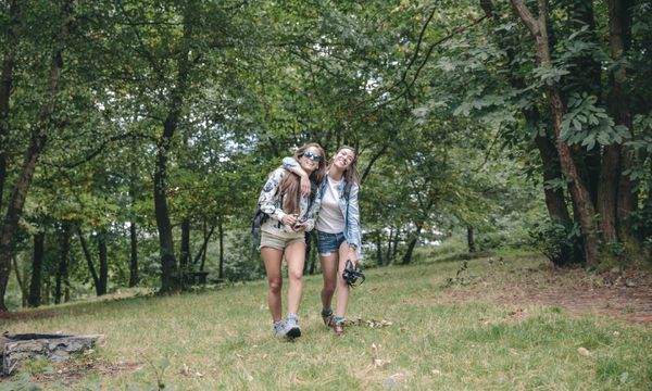 Zwei junge Frauen gehen Arm in Arm durch einen Wald. 