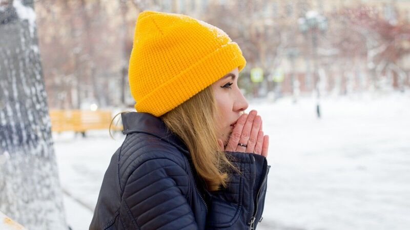 Junge Frau mit gelber Mütze steht in einer winterlichen Landschaft und wärmt sich die Hände.