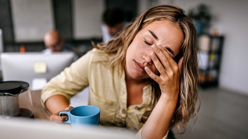 Eine Frau mit Kaffeetasse in einem Büro reibt sich mit geschlossenen Augen das Gesicht