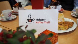 Eine Gruppe verschiedener Personen sitzt an einer Kaffee-Tafel. Eine Helferin der Lohner Malteser reicht Kaffee zum Kuchen.