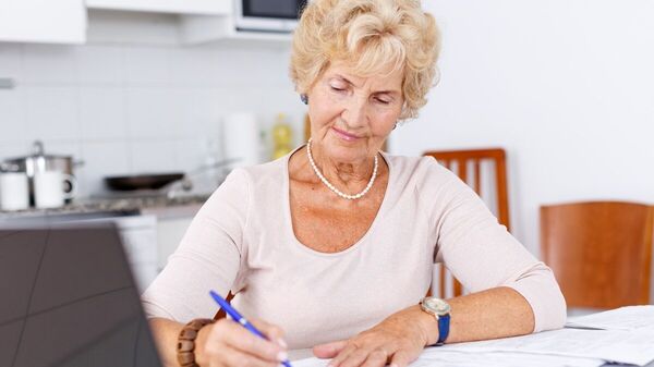 Eine ältere Frau sitzt am Schreibtisch mit einem Laptop und schreibt etwas auf einem Blatt.