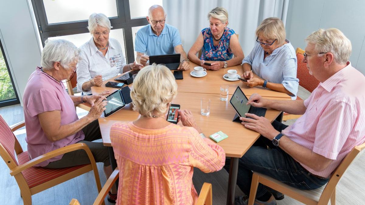 Ältere Menschen mit Kaffeetassen, Tablets und Smartphones an einem Tisch.