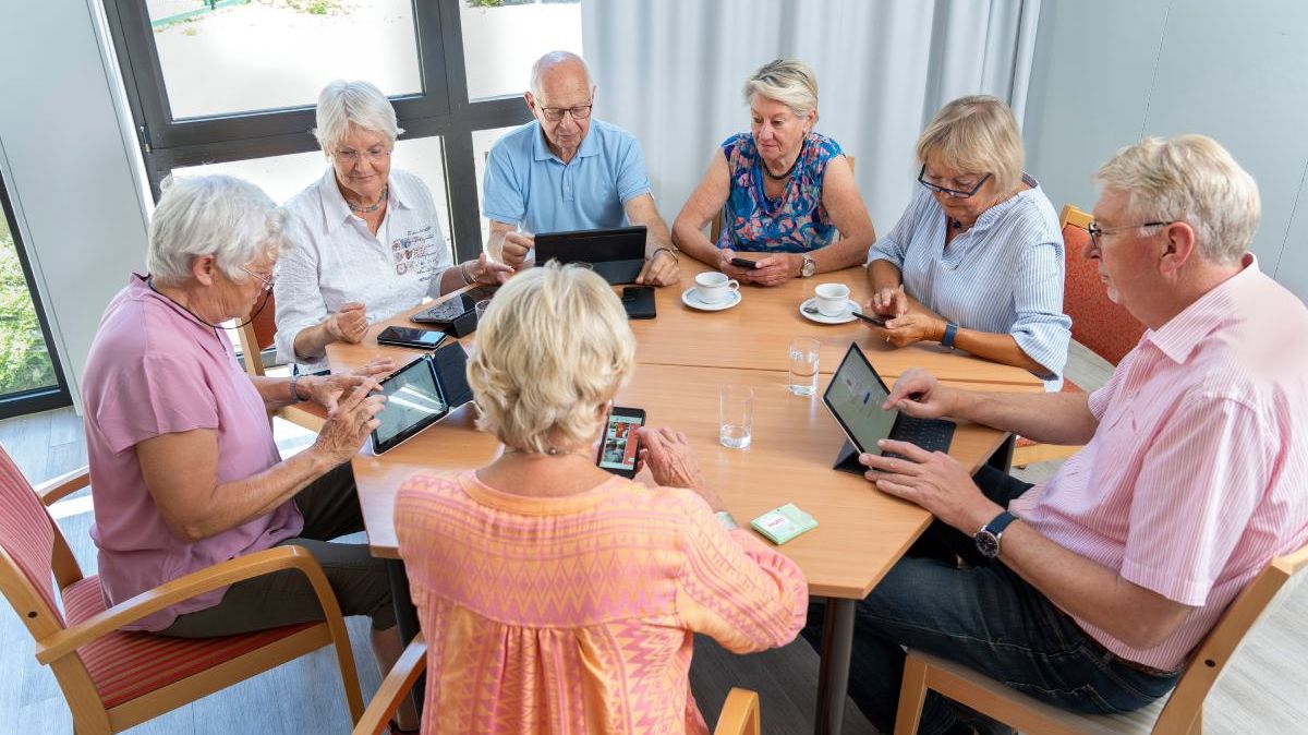 Ältere Menschen mit Kaffeetassen, Tablets und Smartphones an einem Tisch.
