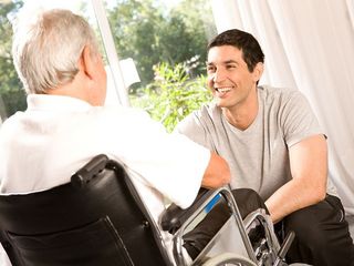 Junger Mann, der vor einer älteren Person im Rollstuhl kniet Malteser