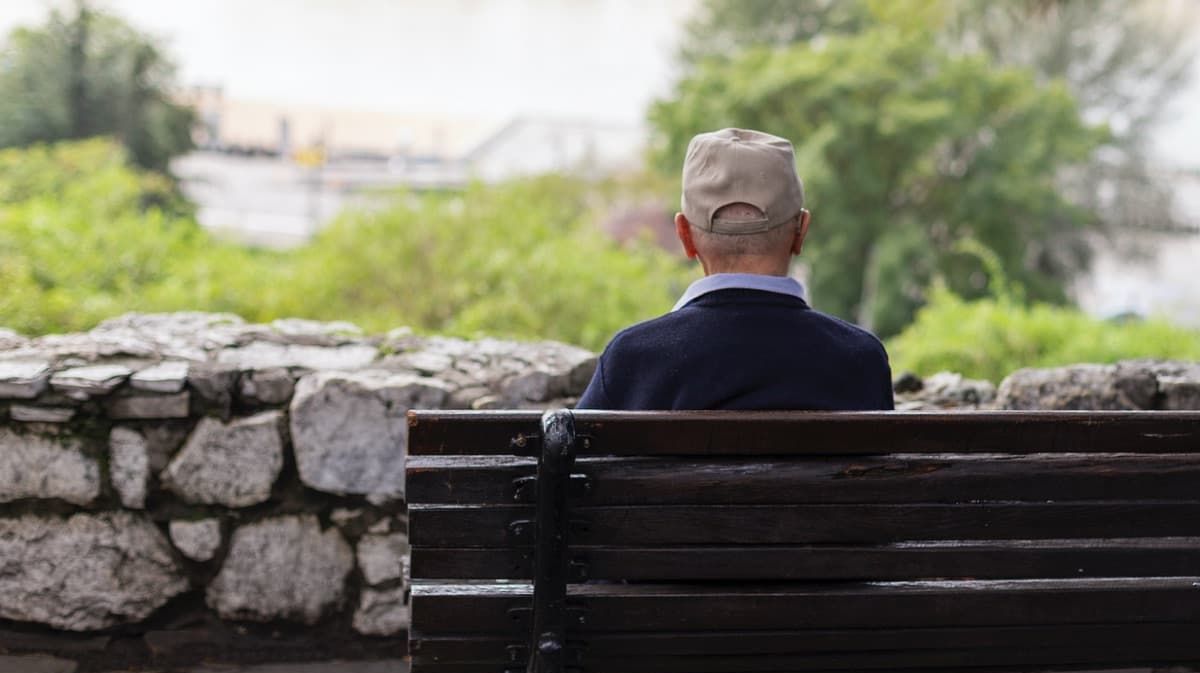 Älterer Mann sitzt mit dem Rücken zur Kamera auf einer Parkbank.