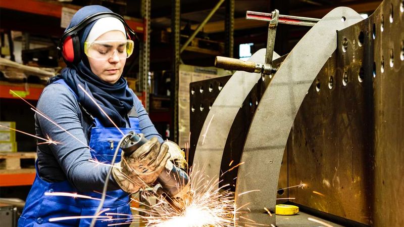 Eine Frau in Arbeitskleidung und mit Kopftuch arbeitet an einem Werkstück aus Metall.