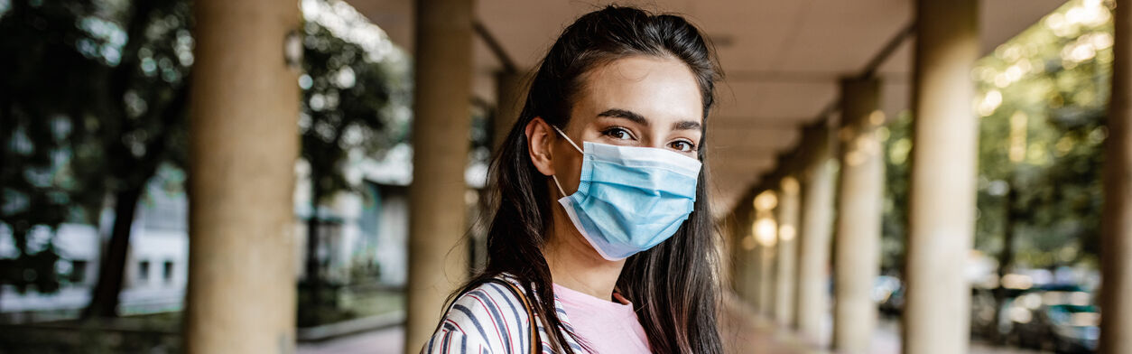 Junge Frau mit Hefter steht mit Mund-Nasen-Schutz auf einem Campus-Gelände.