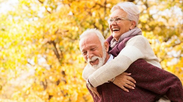 Älterer Mann nimmt eine ältere Frau unter gelbem Herbstlaub Huckepack.