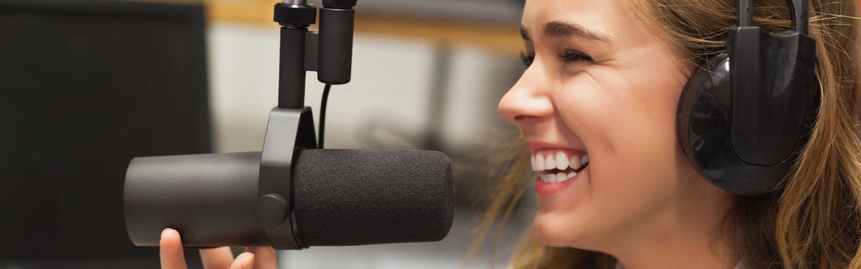 Junge Frau mit Kopfhörern sitzt lächelnd vor einem Mikrofon.