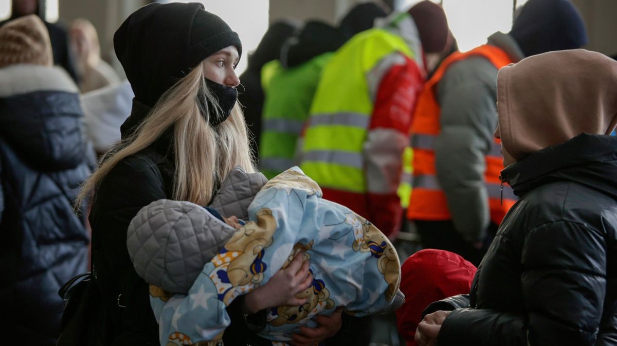 Schwarz gekleidete Frau mit einem Kleinkind im Arm. Im Hintergrund Menschen mit Warnwesten.