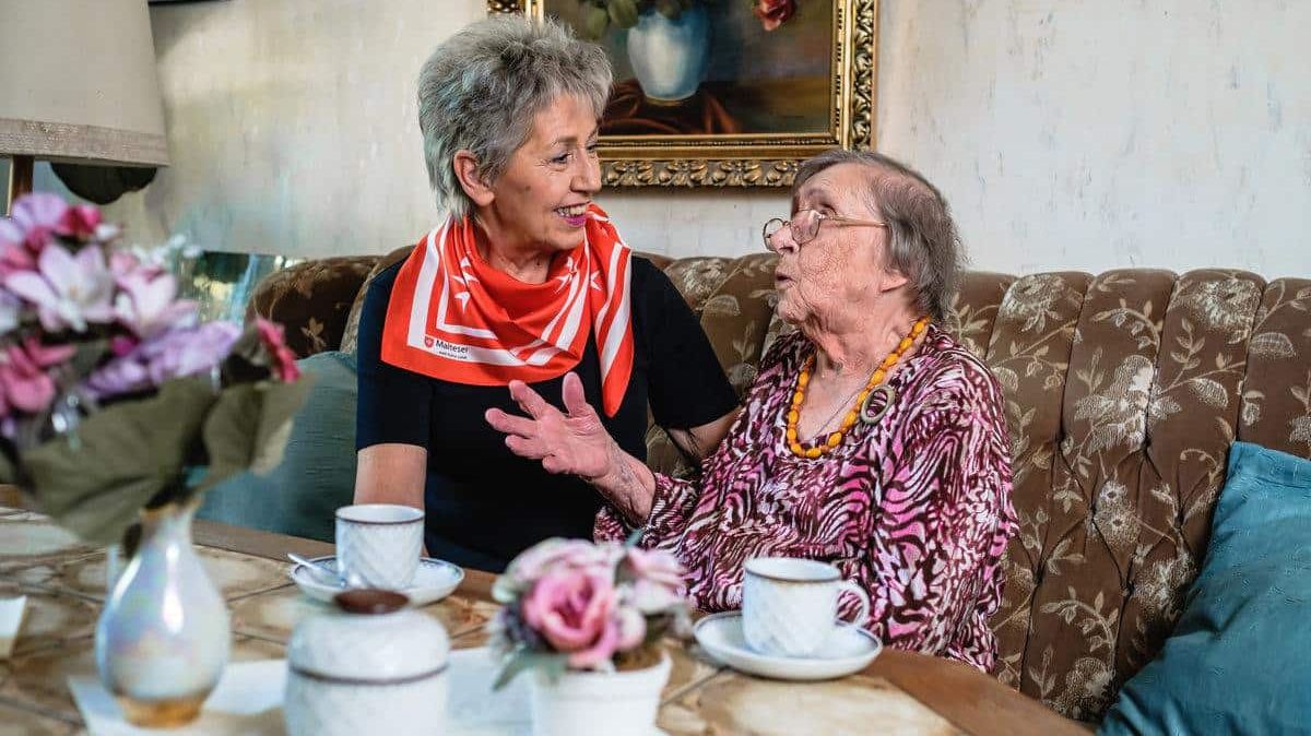 Zwei ältere Frauen sitzen in einem Wohnzimmer auf einem Sofa vor Kaffeetassen.