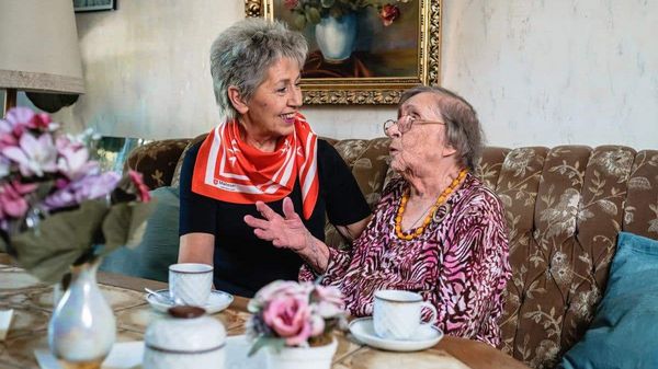 Zwei ältere Frauen sitzen in einem Wohnzimmer auf einem Sofa vor Kaffeetassen.